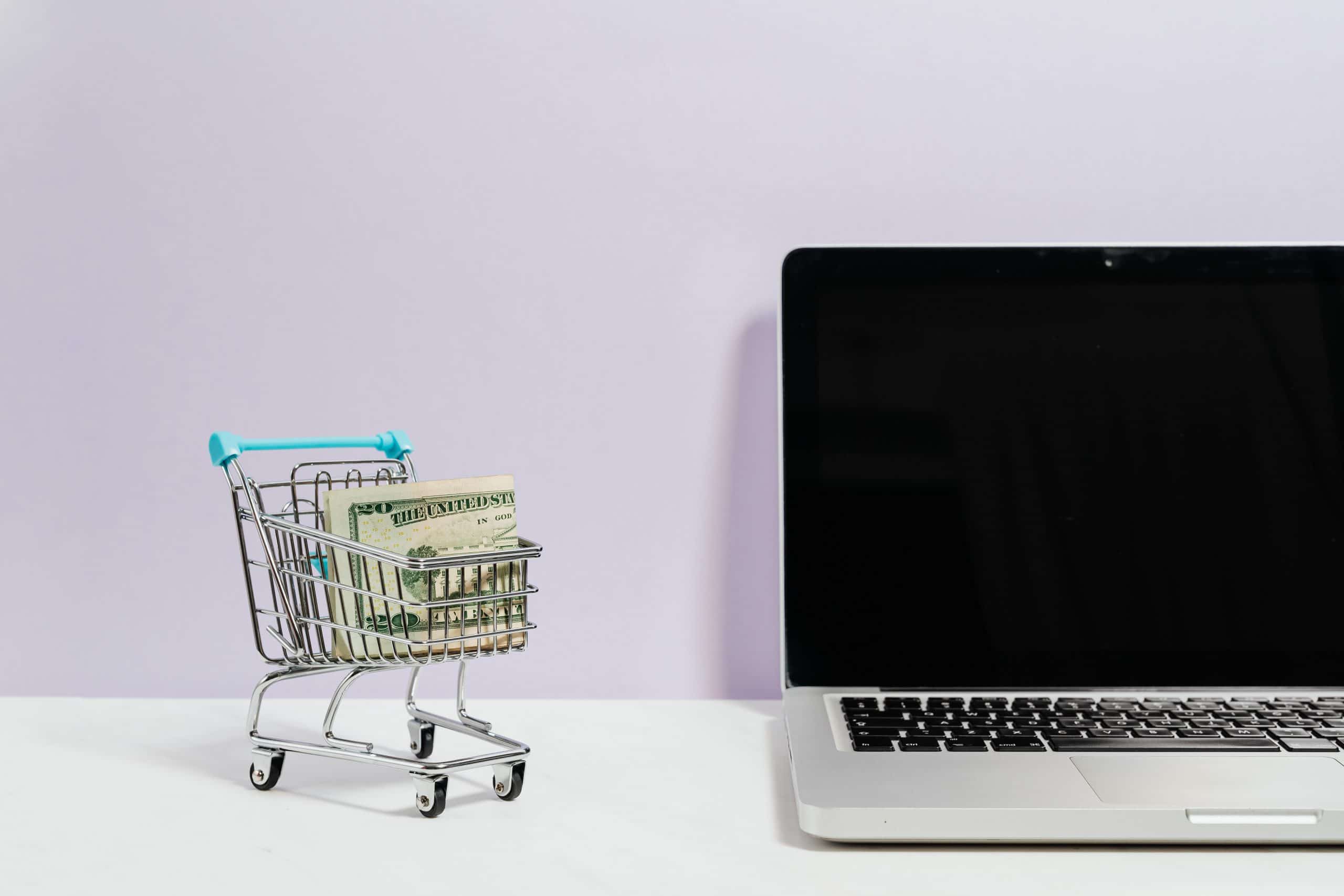 Shopping cart with money next to a laptop computer.