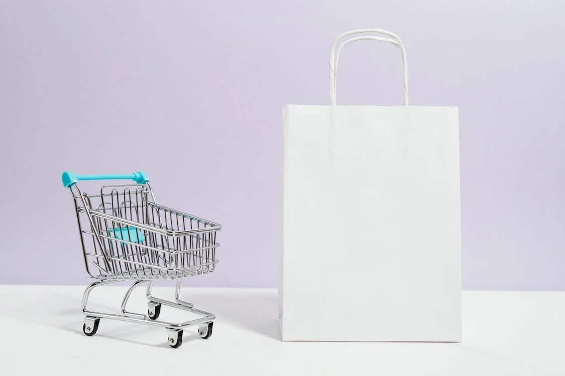 A miniature shopping cart next to a white shopping bag.