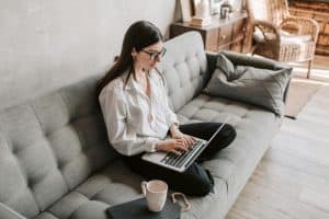 A woman wearing glasses and a white shirt sits cross-legged on a gray sofa, typing on a laptop. A notebook and mug are on the sofa beside her in a cozy, well-lit living room.
