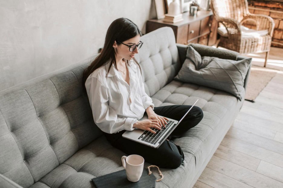 Building Resilience- Self-Care Strategies Every Entrepreneur Needs for Long-Term Success A woman wearing glasses and a white shirt sits cross-legged on a gray sofa, typing on a laptop. A notebook and mug are on the sofa beside her in a cozy, well-lit living room.