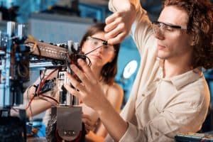 Two people wearing safety goggles work on assembling or repairing a robot, using tools and connecting wires in a workshop or laboratory setting.