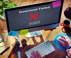 A computer screen displays a Download Failed message with a red X and a try again button. A person uses a mouse at a cluttered desk with papers, sticky notes, coffee, and office supplies.