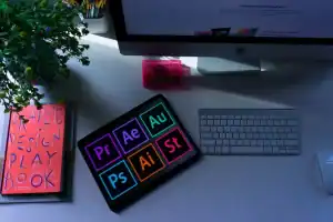 Tablet displaying Adobe Creative Cloud app icons beside a design desk setup with iMac and keyboard