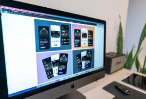 A computer monitor displays a design software window showing multiple mobile wallet app interface mockups in various colors, each demonstrating elements of responsive design. A plant, speaker, keyboard, and mouse are on the desk nearby.