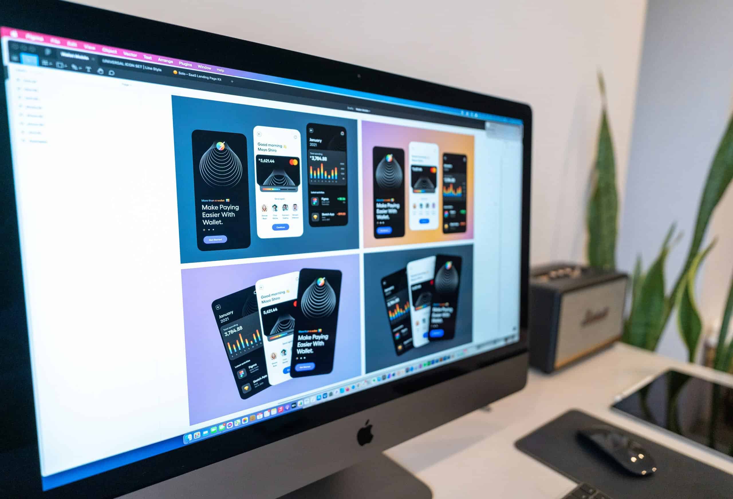 photo-1609921212029-bb5a28e60960 A computer monitor displays a design software window showing multiple mobile wallet app interface mockups in various colors, each demonstrating elements of responsive design. A plant, speaker, keyboard, and mouse are on the desk nearby.